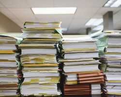 Stack of more than twenty binders filled with hundreds of papers on a table in an office space.