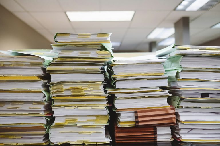 Stack of more than twenty binders filled with hundreds of papers on a table in an office space.